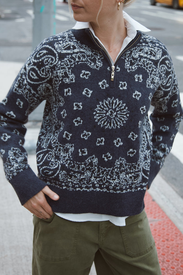 A person wearing a navy quarter-zip sweater with a white paisley and floral bandana pattern, styled with a white collared shirt and olive green pants on an outdoor street setting.