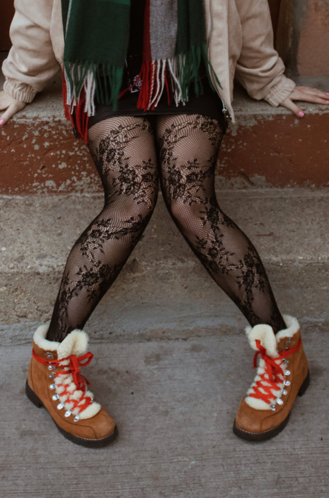 Person wearing a plaid scarf, beige jacket, black patterned tights, and brown boots on stone steps.