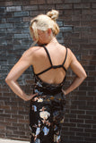 A woman with blonde hair in a bun is shown from behind, wearing a black strappy dress with a floral print, posing against a dark brick wall.