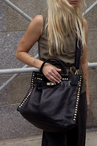 A woman wearing a sleeveless olive green top and black pants carries a large black leather handbag with gold-tone studs and hardware. The photo is taken outdoors against a gray stone wall and metal scaffolding.