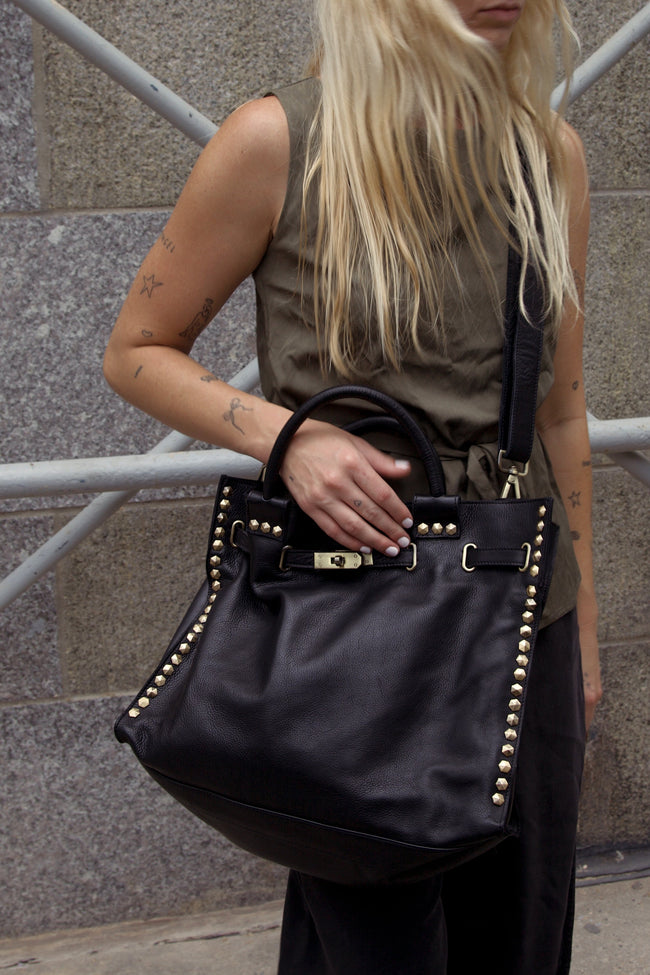 A woman wearing a sleeveless olive green top and black pants carries a large black leather handbag with gold-tone studs and hardware. The photo is taken outdoors against a gray stone wall and metal scaffolding.