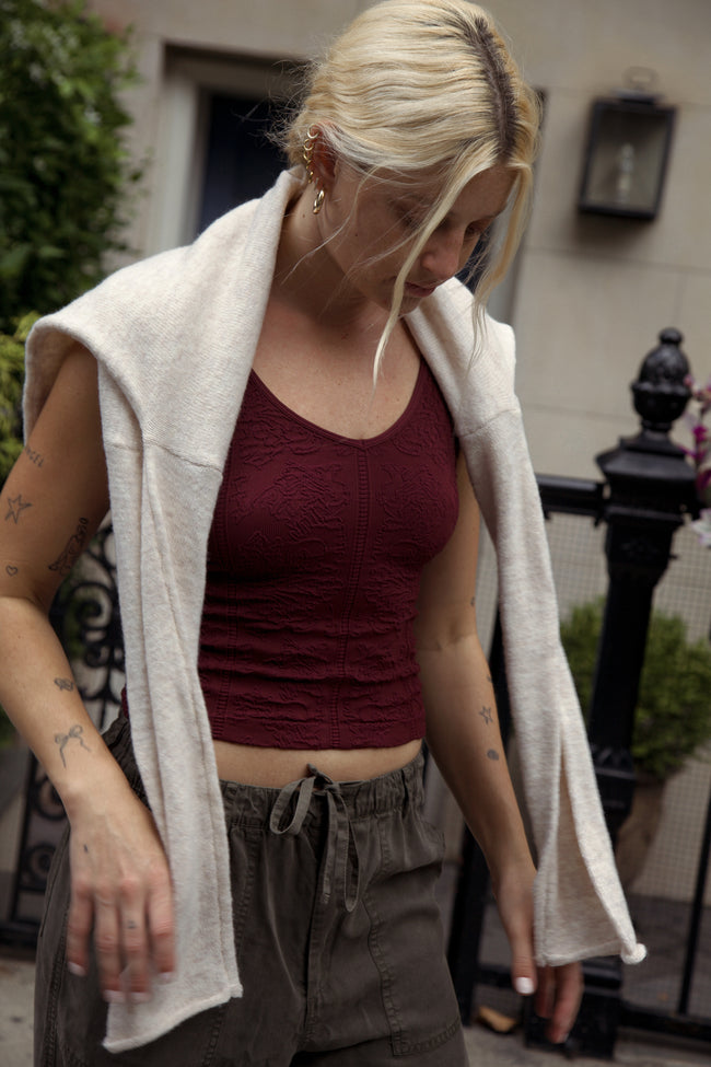 A woman wearing a textured burgundy ribbed tank top, a cream knit cardigan draped off her shoulders, and olive green drawstring pants, standing outdoors in front of a building.