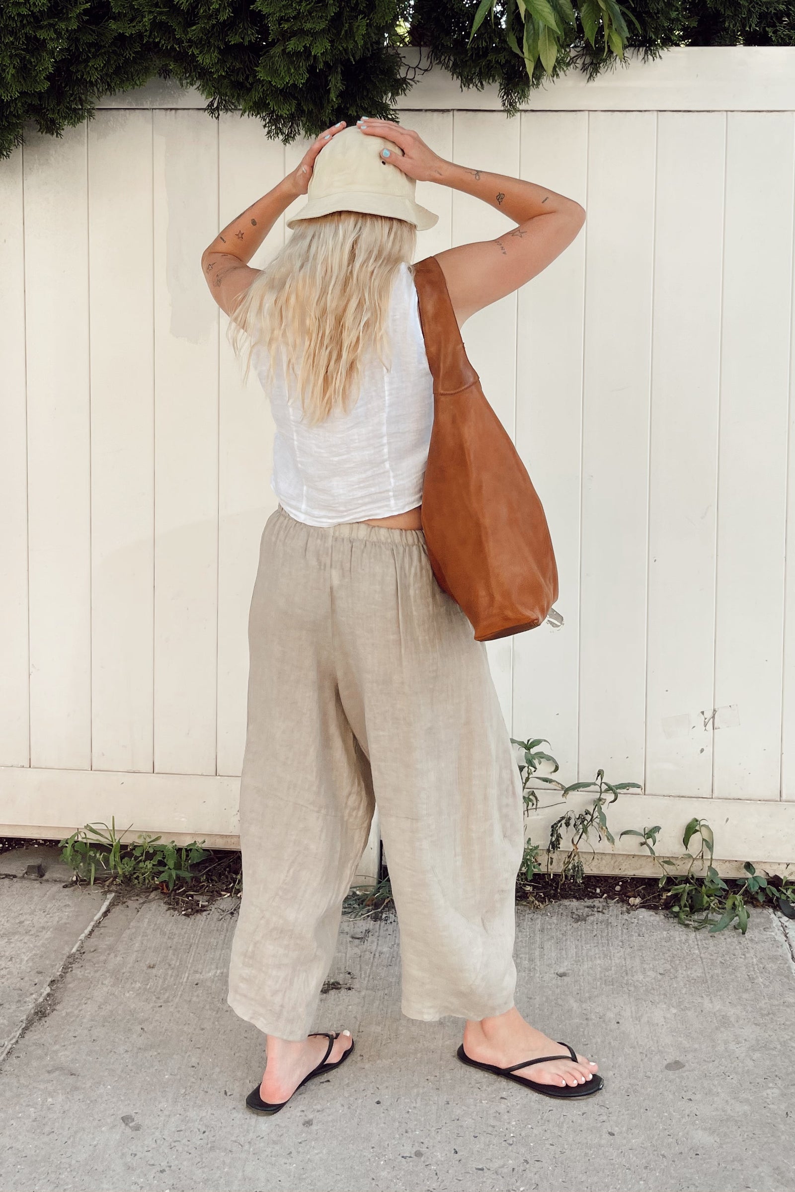 A woman stands with her back to the camera wearing a white sleeveless top, beige wide-leg linen pants, a cream bucket hat, and black flip-flops, while carrying a large brown leather shoulder bag against a white wooden fence.
