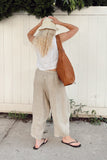 A woman stands with her back to the camera wearing a white sleeveless top, beige wide-leg linen pants, a cream bucket hat, and black flip-flops, while carrying a large brown leather shoulder bag against a white wooden fence.