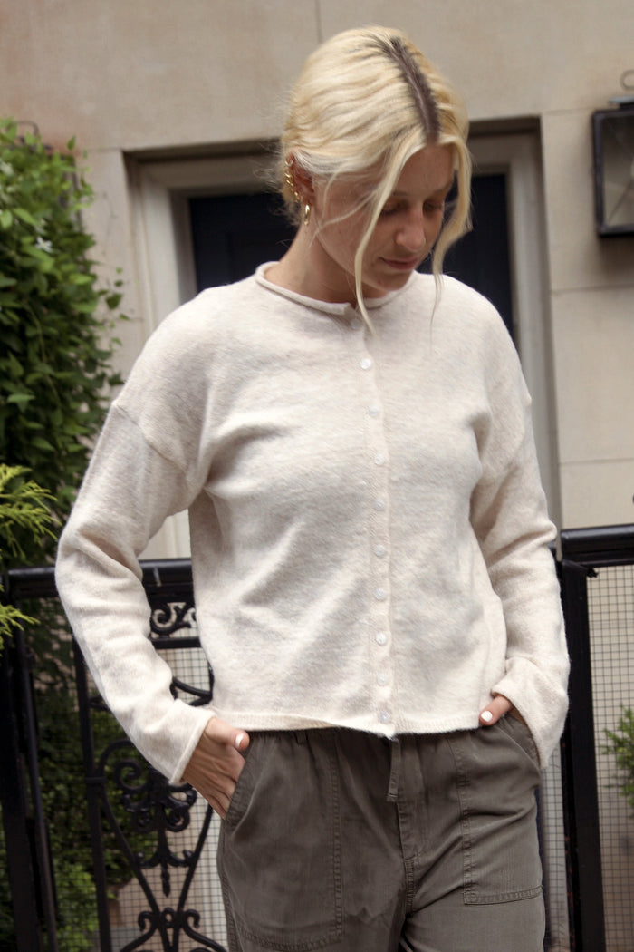A woman stands outdoors wearing a cream knit cardigan, wide-leg olive pants, and black flats, with a black wrought-iron fence and building facade behind her.