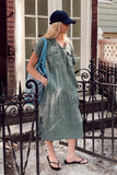 A woman wearing a sage green linen midi dress, a navy blue baseball cap, and black flip-flops stands on a porch in front of a black wrought-iron gate. She is carrying a blue patterned shoulder bag.