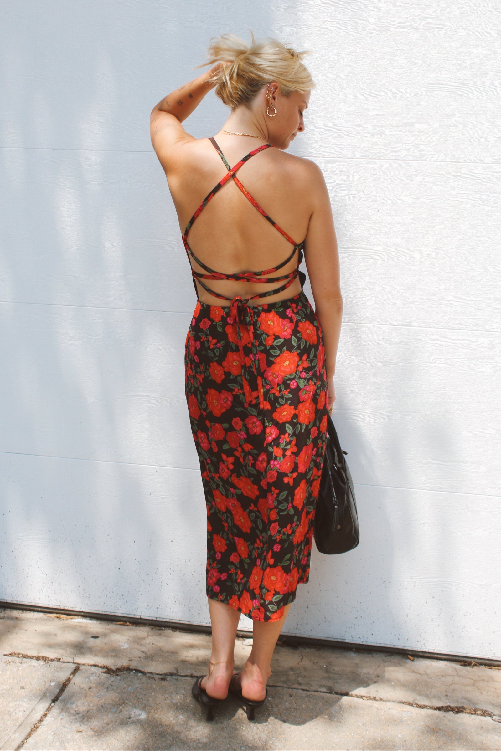 A woman stands with her back to the camera wearing a red and black floral midi dress with a strappy open-back design, holding a black handbag against a white wall.