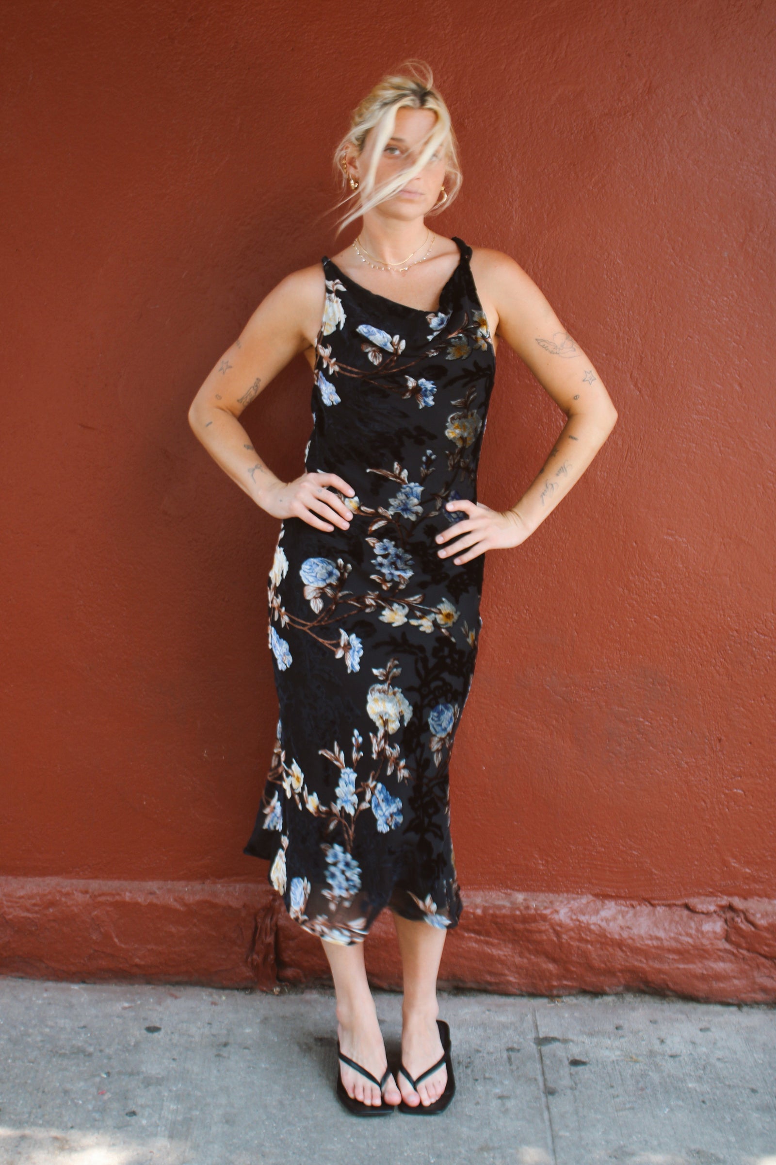 A woman wearing a black midi dress with a blue and white floral pattern, standing against a textured reddish-brown wall.
