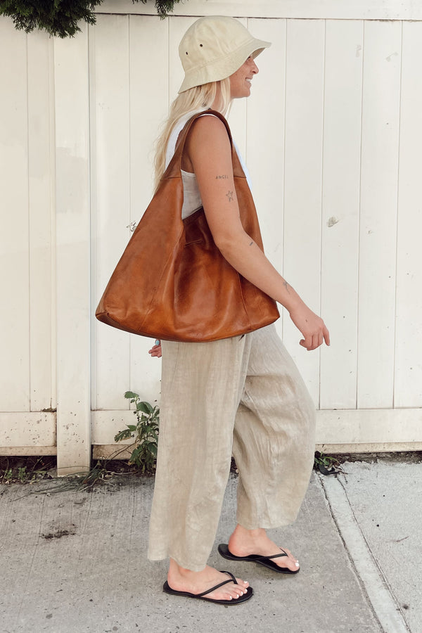 A woman wearing a cream bucket hat, a white sleeveless top, beige wide-leg linen pants, and black flip-flops carries a large brown leather tote bag as she walks in front of a white wooden fence.