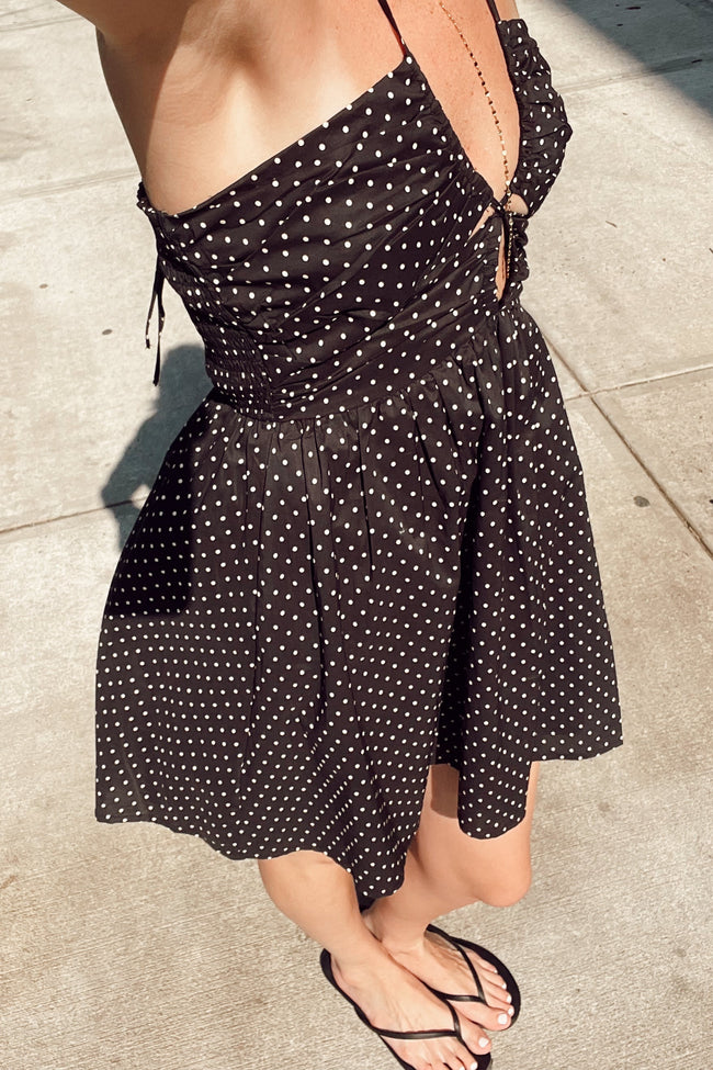 A woman wearing a black spaghetti-strap dress with a white polka dot pattern, styled with black flip-flops on a concrete sidewalk.