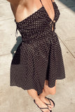 A woman wearing a black spaghetti-strap dress with a white polka dot pattern, styled with black flip-flops on a concrete sidewalk.
