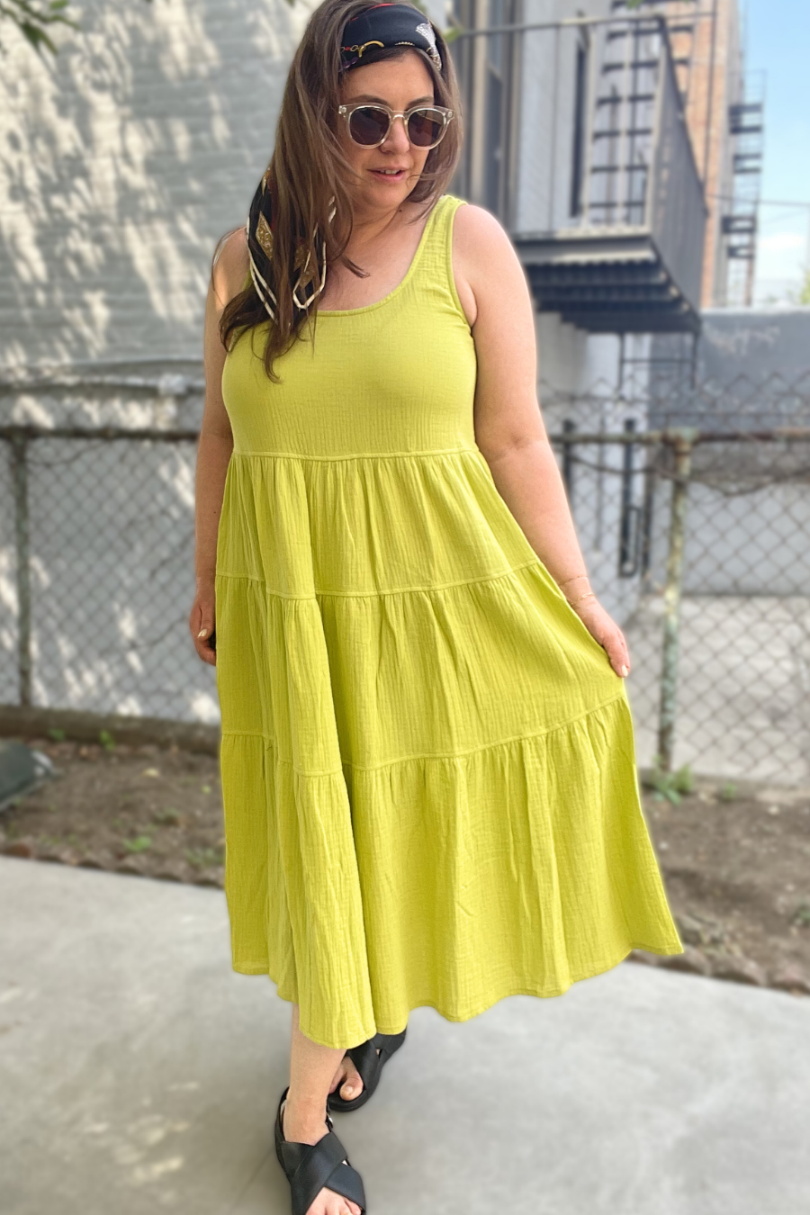 Woman wearing a yellow dress standing outdoors with a chain-link fence and building in the background.