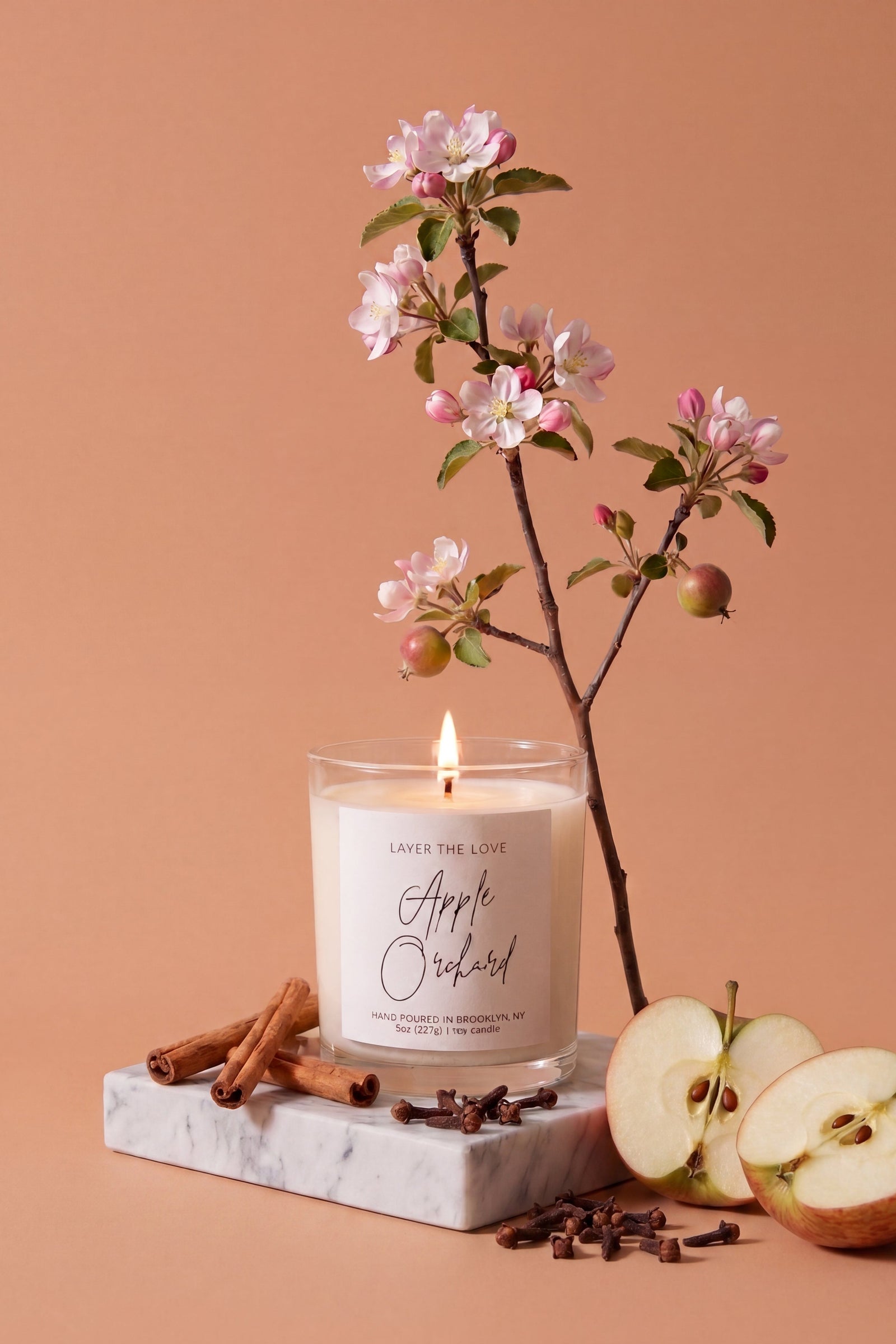 A lit scented candle in a clear glass jar sits on a white marble block, surrounded by apple slices, cinnamon sticks, cloves, and a flowering branch against a warm peach background. The candle label reads 'Layer the Love Apple Orchard'.