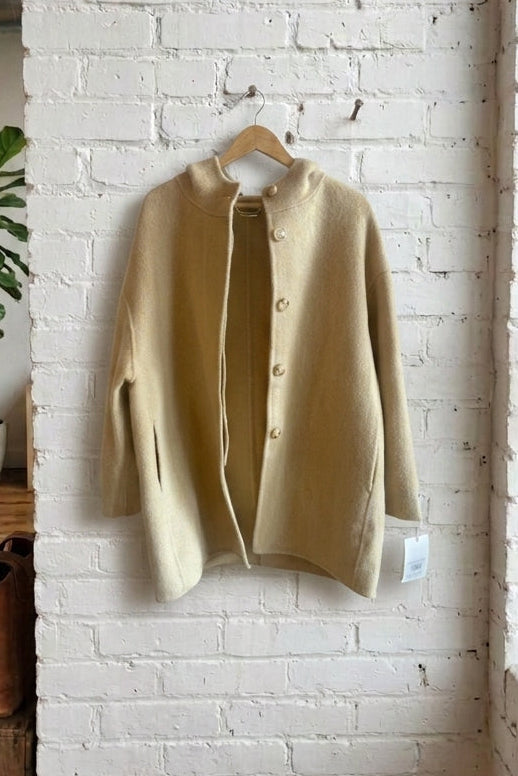A light beige knit coat hanging on a wooden hanger against a white brick wall in a loft-style room with a window, wooden crate, and potted plant.