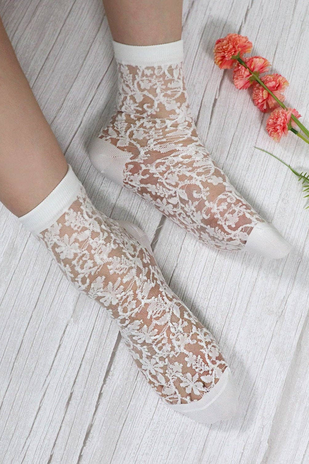 White lace ankle socks worn on feet, shown on a light wood floor with decorative flowers in the corner.