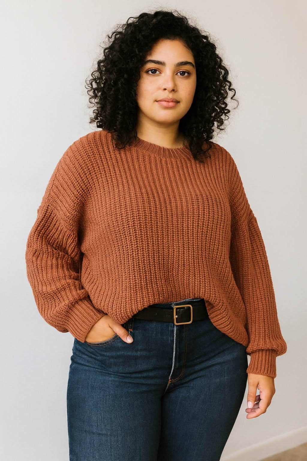 A woman with curly black hair wearing a rust-brown ribbed knit sweater and dark blue jeans with a black belt, standing against a plain off-white wall.