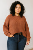 A woman with curly black hair wearing a rust-brown ribbed knit sweater and dark blue jeans with a black belt, standing against a plain off-white wall.