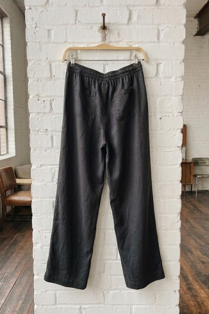 Black wide-leg tencel trousers hanging on a wooden hanger from a brass hook on a white brick pillar in a loft-style room with wooden floors.