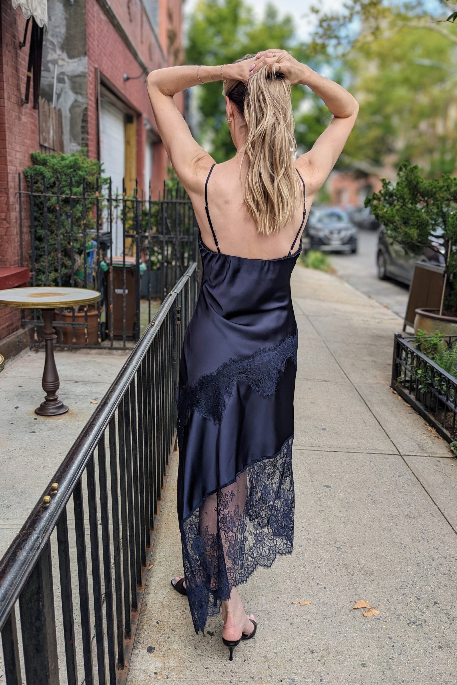A woman wearing a dark navy blue satin slip dress with black lace hem, standing on a city sidewalk and looking over her shoulder.