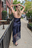 A woman wearing a dark navy blue satin slip dress with black lace hem, standing on a city sidewalk and looking over her shoulder.
