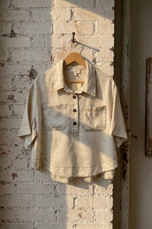A light beige short-sleeve button-up shirt with two chest pockets, hanging on a wooden hanger against a white brick wall with warm sunlight and shadows.