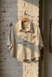 A light beige short-sleeve button-up shirt with two chest pockets, hanging on a wooden hanger against a white brick wall with warm sunlight and shadows.