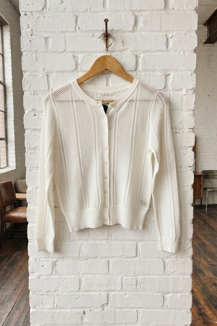 White knit cardigan on a wooden hanger against a white brick wall.