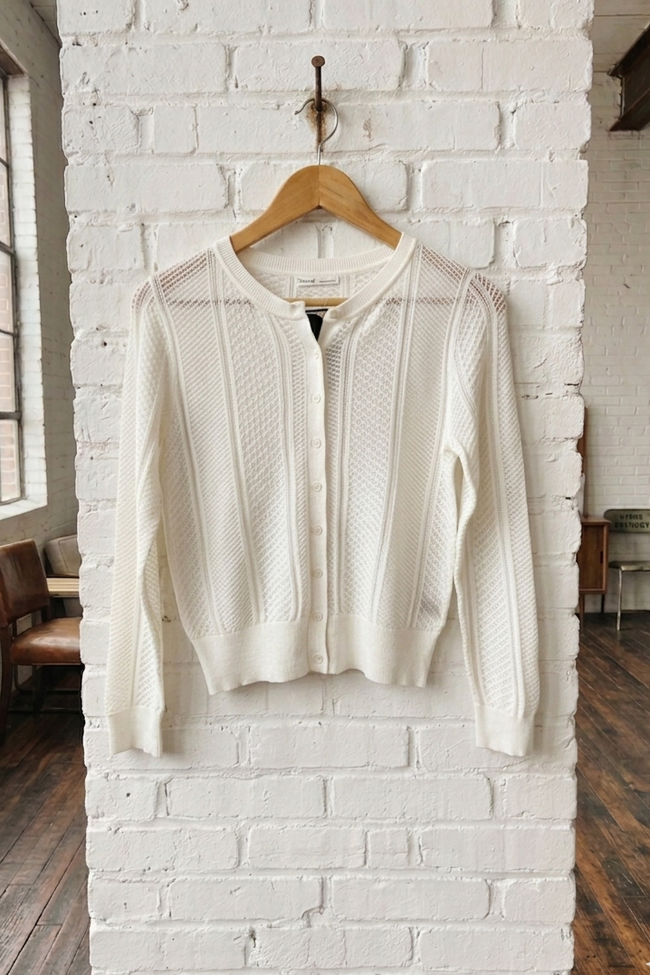 White knit cardigan on a wooden hanger against a white brick wall.
