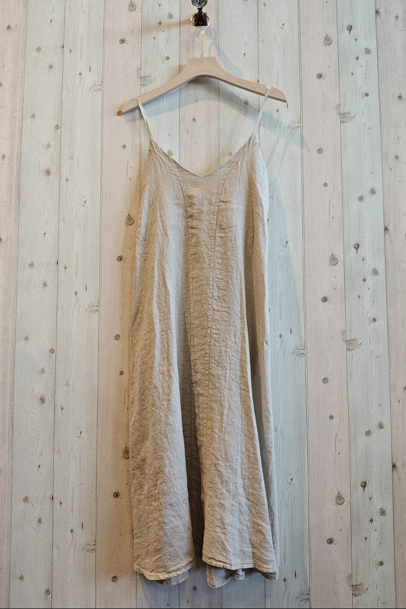 A light beige linen slip dress with thin spaghetti straps hanging from a wooden hanger against a light wood plank wall.