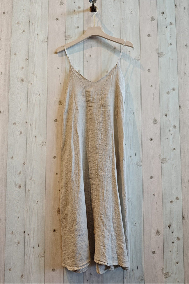 A light beige linen slip dress with thin spaghetti straps hanging from a wooden hanger against a light wood plank wall.