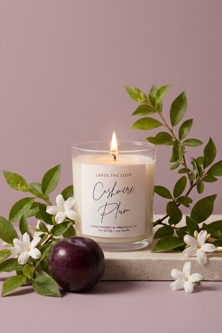 A lit candle in a clear glass jar sits on a stone slab surrounded by green leaves, white flowers, and a dark purple plum against a dusty mauve background. The candle label reads 'Layer the Love Cinnamon Plum'.