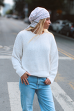 A woman standing on a city street crosswalk wearing a white ribbed knit sweater, light blue wide-leg jeans, white shoes, and a light patterned headscarf.