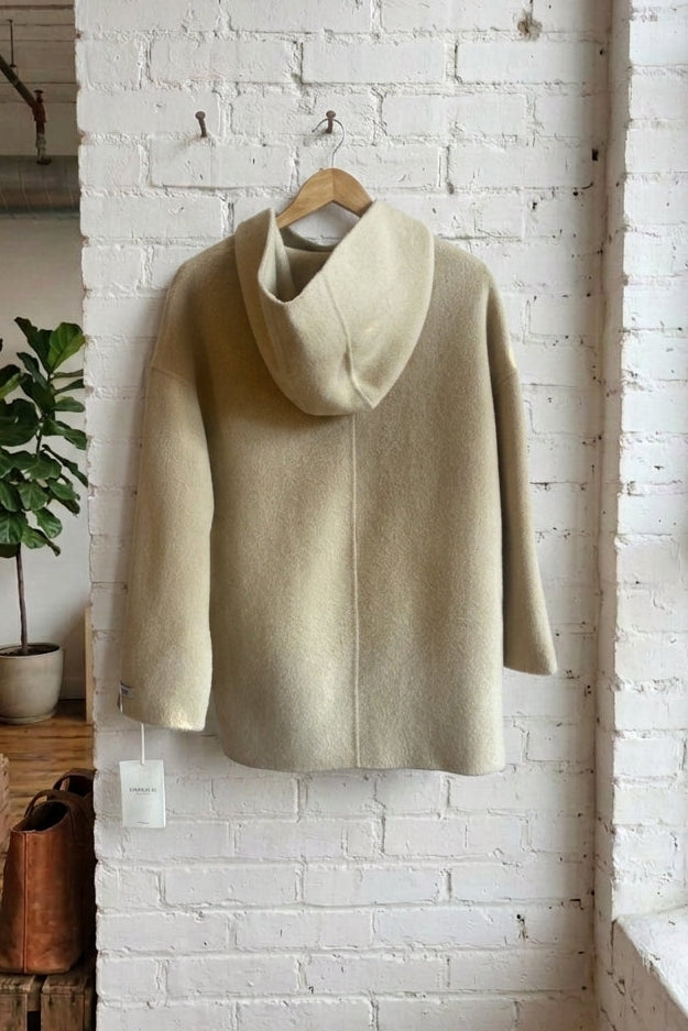 Back view of a light beige coat with a hood and long sleeves, hanging on a wooden hanger against a white brick wall in a loft-style room with a window and potted plant.