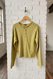 A light dijon colored knit cardigan sweater hanging on a wooden hanger against a white brick wall in a loft-style room with dark wood floors.