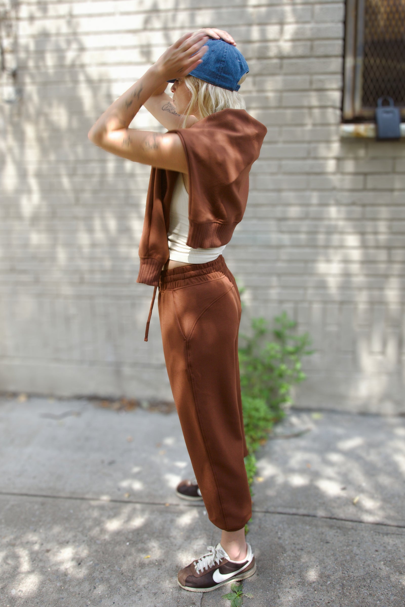A person standing outdoors on a sidewalk in front of a light gray brick wall, wearing a matching brown cropped zip hoodie and jogger pants set, a white tank top, a blue beanie, and brown sneakers.