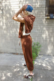 A person standing outdoors on a sidewalk in front of a light gray brick wall, wearing a matching brown cropped zip hoodie and jogger pants set, a white tank top, a blue beanie, and brown sneakers.