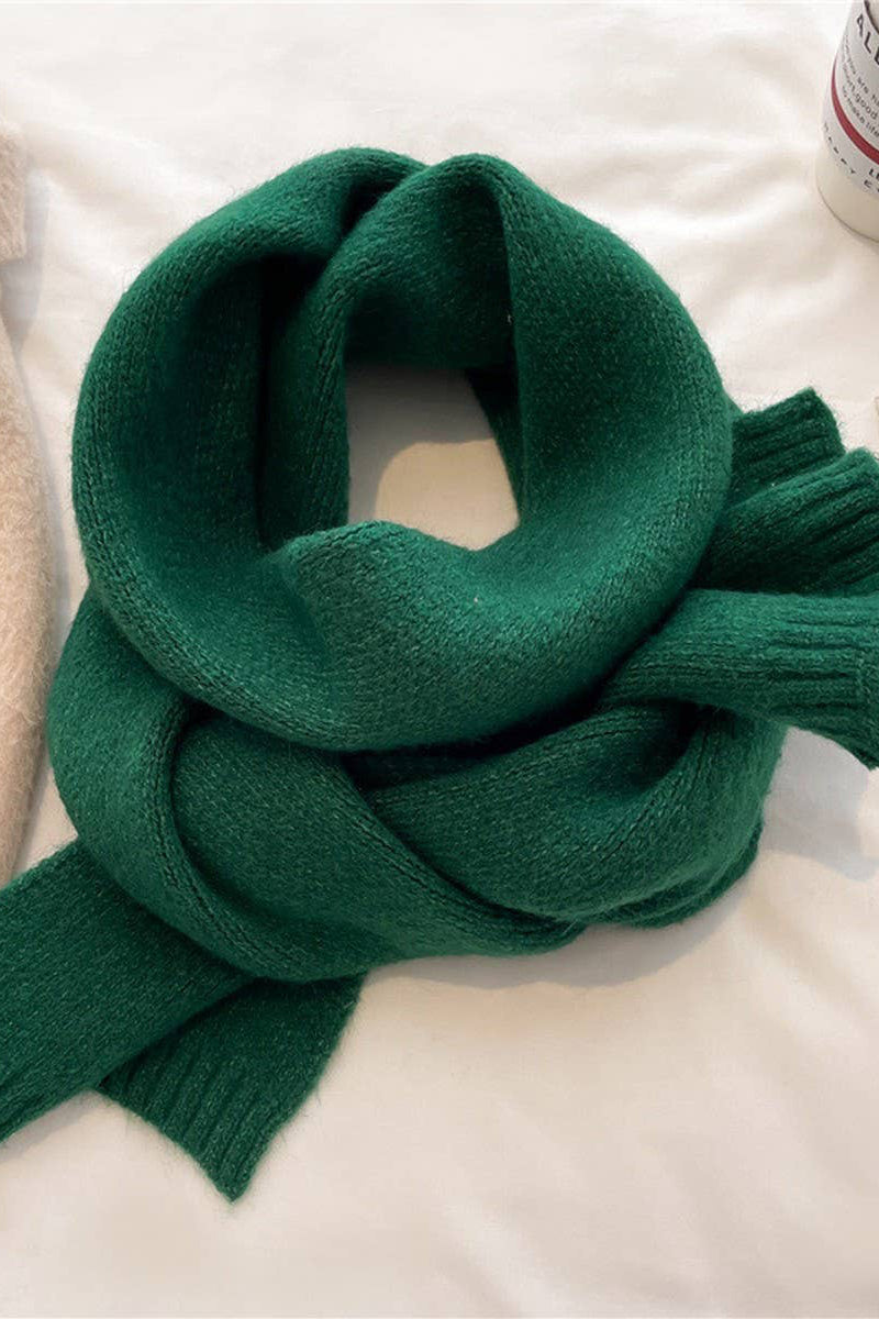 Green knitted scarf on a light surface with a beige garment and magazine in the background