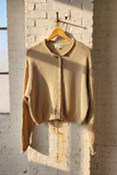 A beige knit cropped cardigan sweater with a button-down front, hanging on a wooden hanger against a white brick wall with warm sunlight and window shadows.