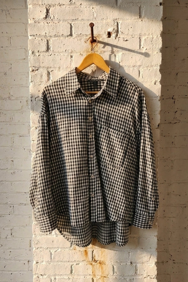A black and white gingham long-sleeve button-up shirt hanging on a wooden hanger against a white brick wall with warm sunlight.