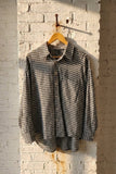 A black and white gingham long-sleeve button-up shirt hanging on a wooden hanger against a white brick wall with warm sunlight.