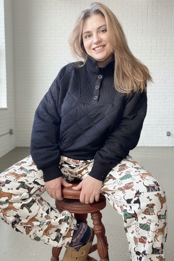 Person wearing a dark sweater and patterned pants sitting on a stool indoors.