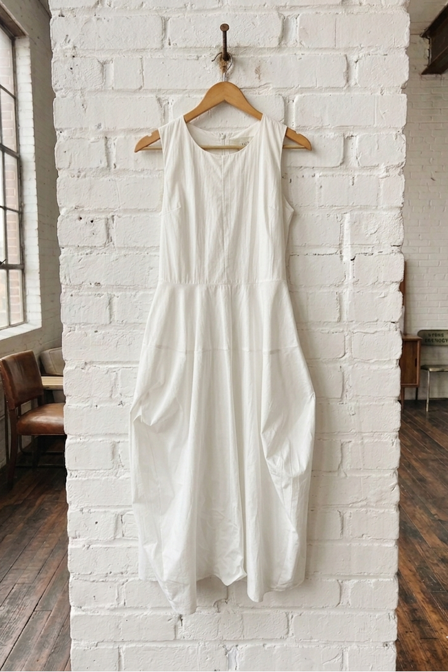 White dress hanging on a wooden hanger against a white brick wall.