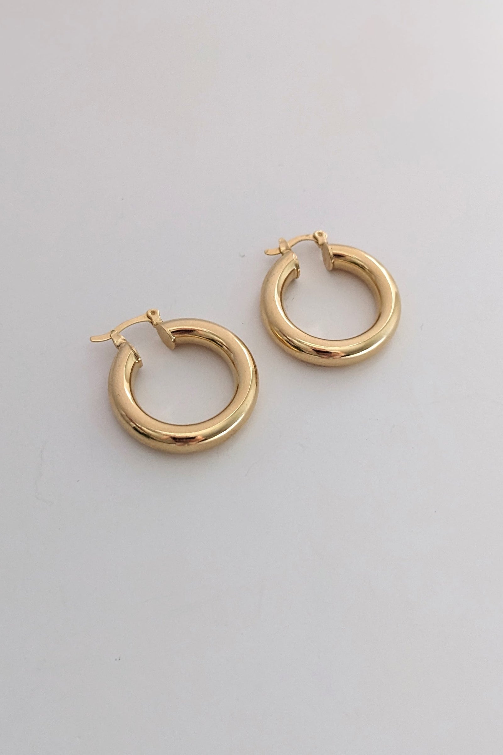 A pair of polished gold-tone hoop earrings with a thick, rounded tubular design and post-back closures, displayed on a clean, light gray surface.