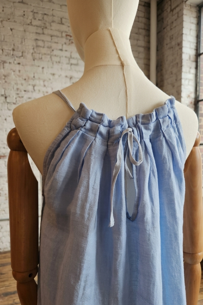 A light blue sleeveless camisole top displayed on a cream mannequin torso, set in a rustic room with a brick wall and large window.