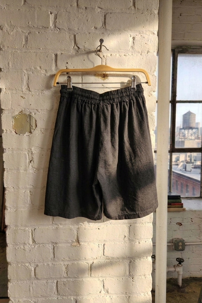 black linen shorts hanging on a hanger against a white brick wall with a cityscape view.