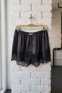 Black satin shorts with a lace hem, hanging on a wooden hanger against a white brick wall.