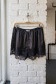 Black satin shorts with a lace hem, hanging on a wooden hanger against a white brick wall.