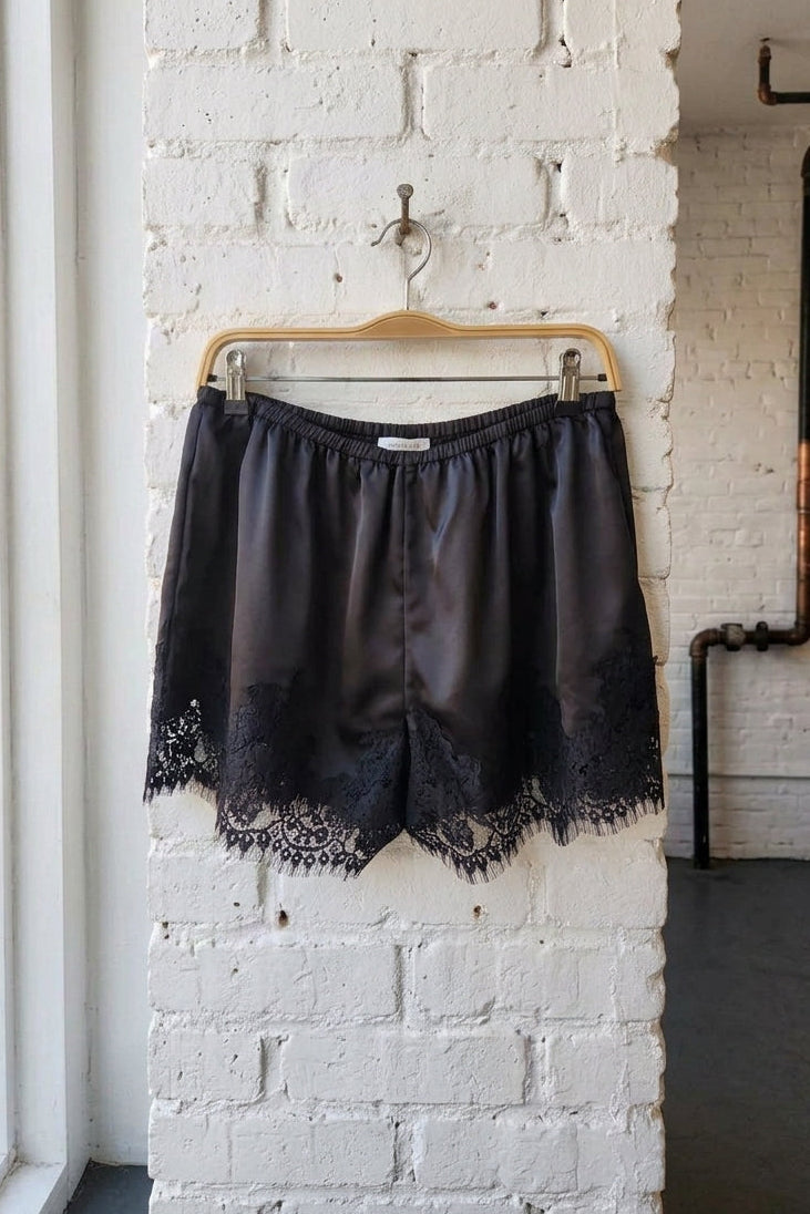 Black satin shorts with a lace hem, hanging on a wooden hanger against a white brick wall.