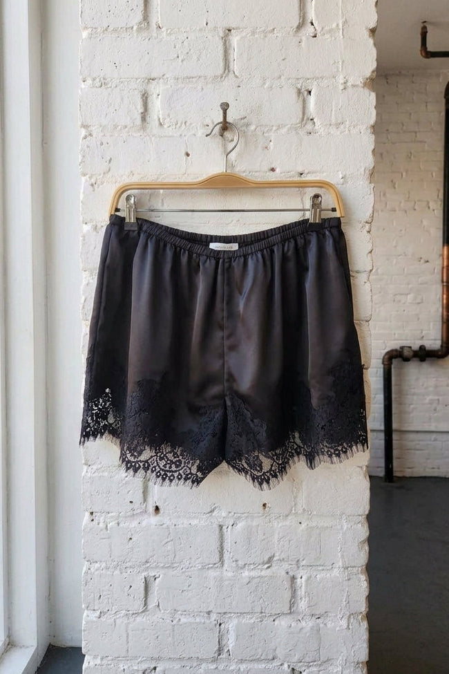Black satin shorts with a lace hem, hanging on a wooden hanger against a white brick wall.