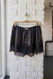 Black satin shorts with a lace hem, hanging on a wooden hanger against a white brick wall.